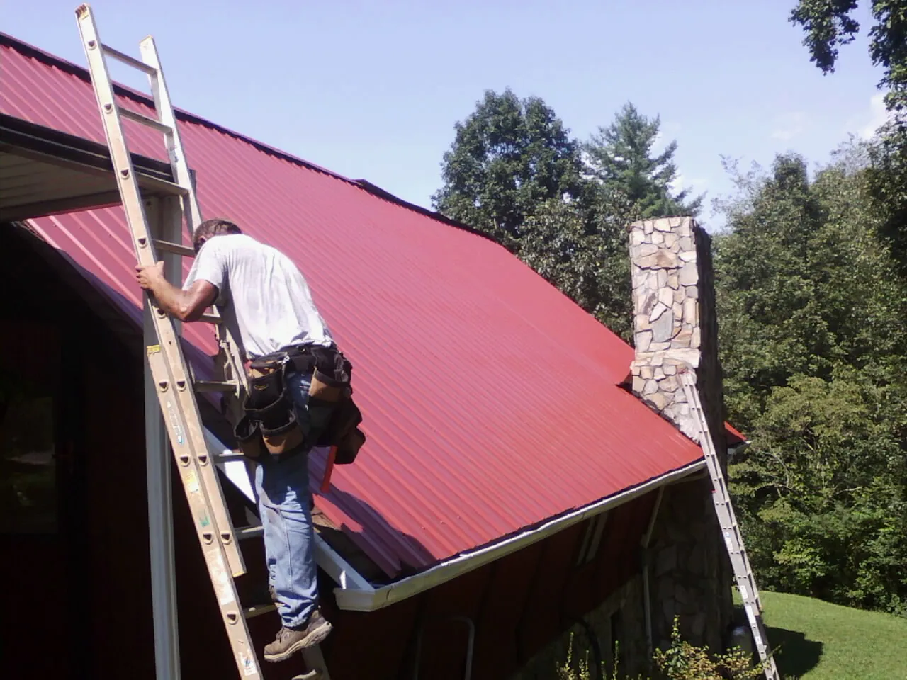 Roofer on a ladder beside a red metal roof and stone chimney — Soffit & Fascia Repair by Roofing Brothers in Bryans Road